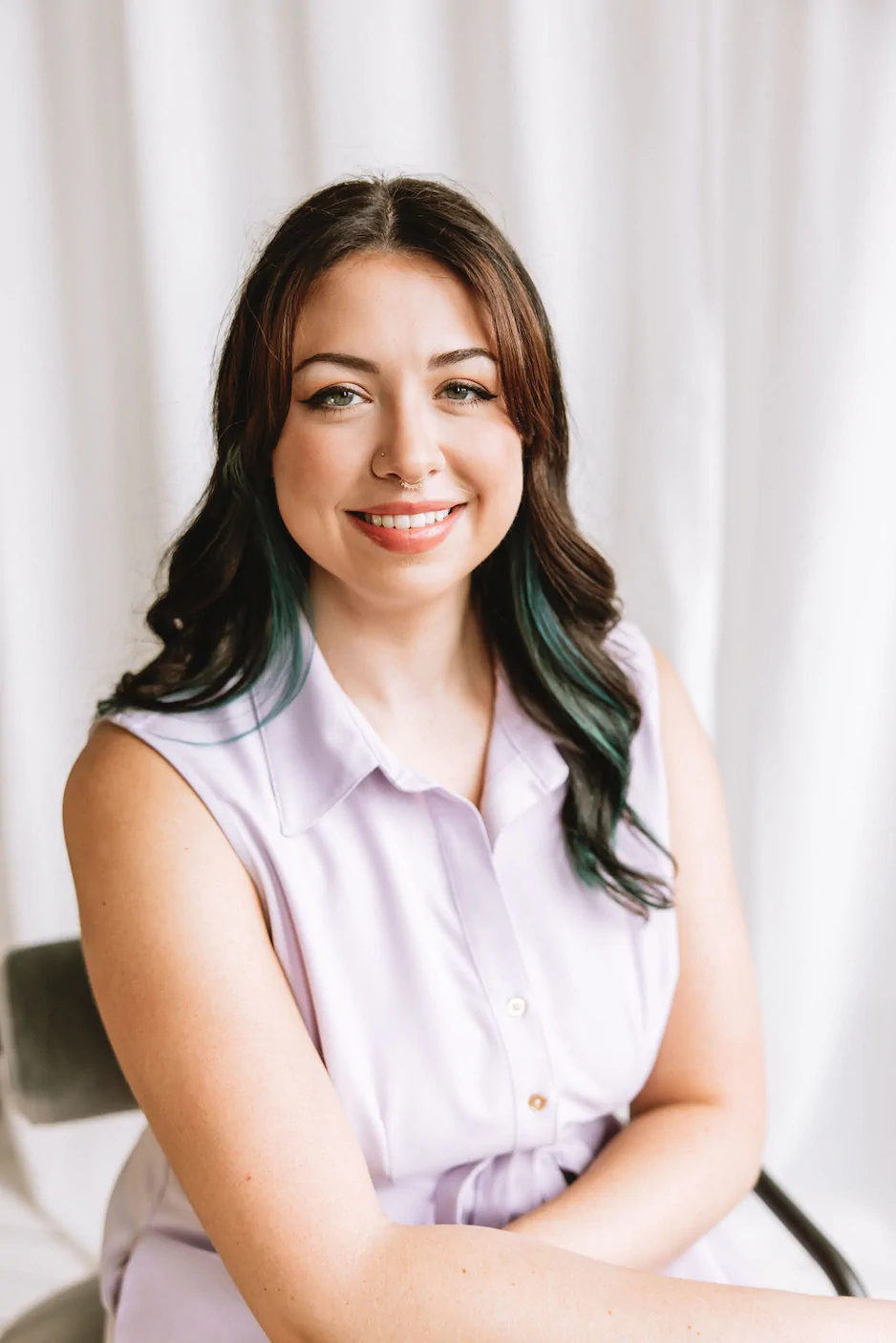 Dietitian Rachel Green is a woman with long brown hair and green highlights sits on a chair, smiling, wearing a sleeveless light purple blouse, with a white curtain background. Ohio