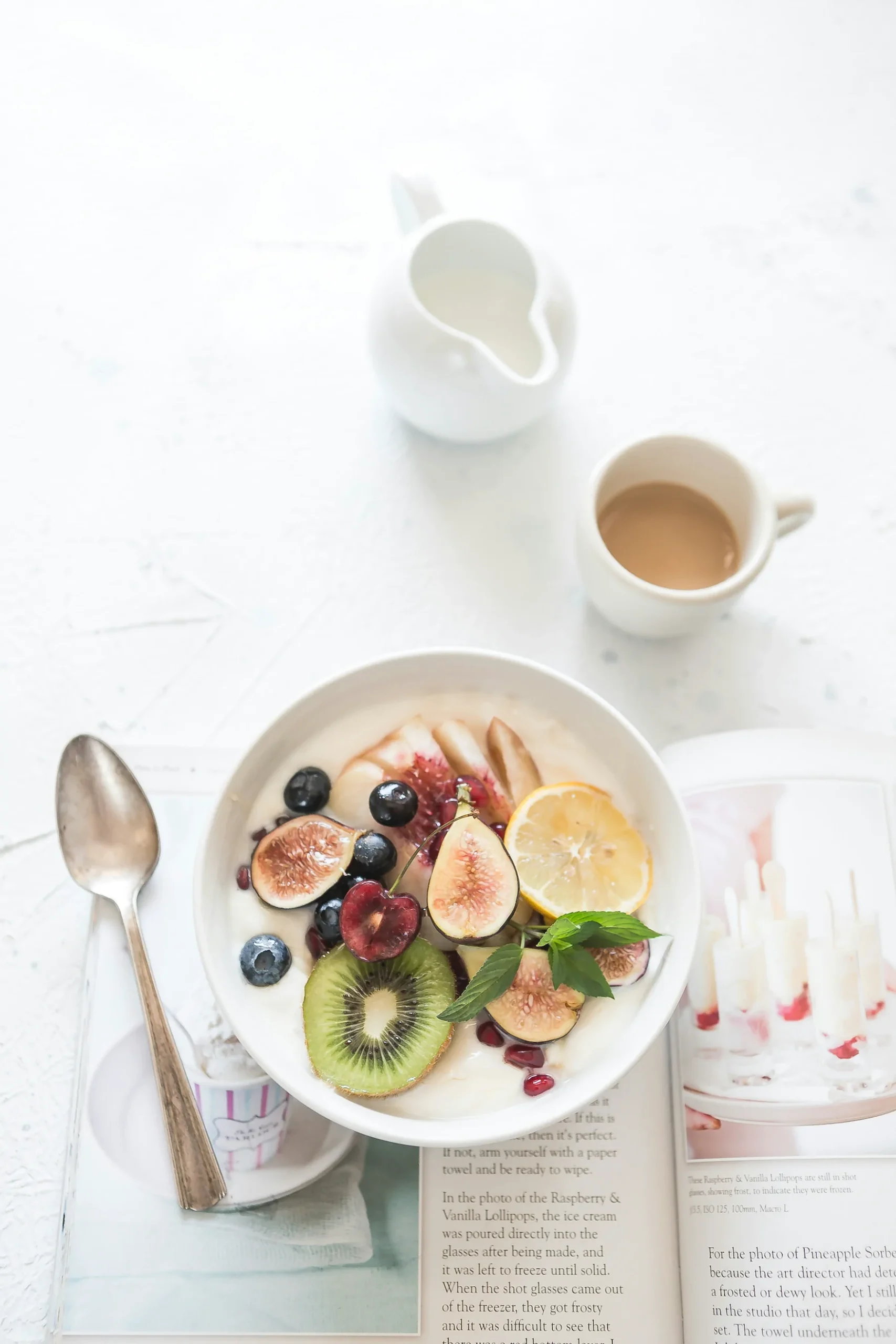 A bowl of fruit and yogurt sits on an open magazine next to a spoon, a cup of coffee, and a small white pitcher on a white surface. Ohio