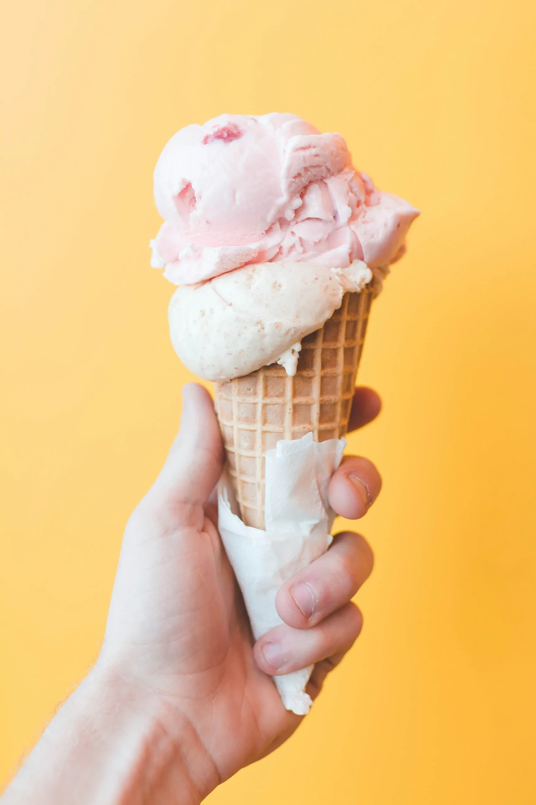 A hand holding a waffle cone with two scoops of pink and white ice cream against a solid yellow background. Ohio