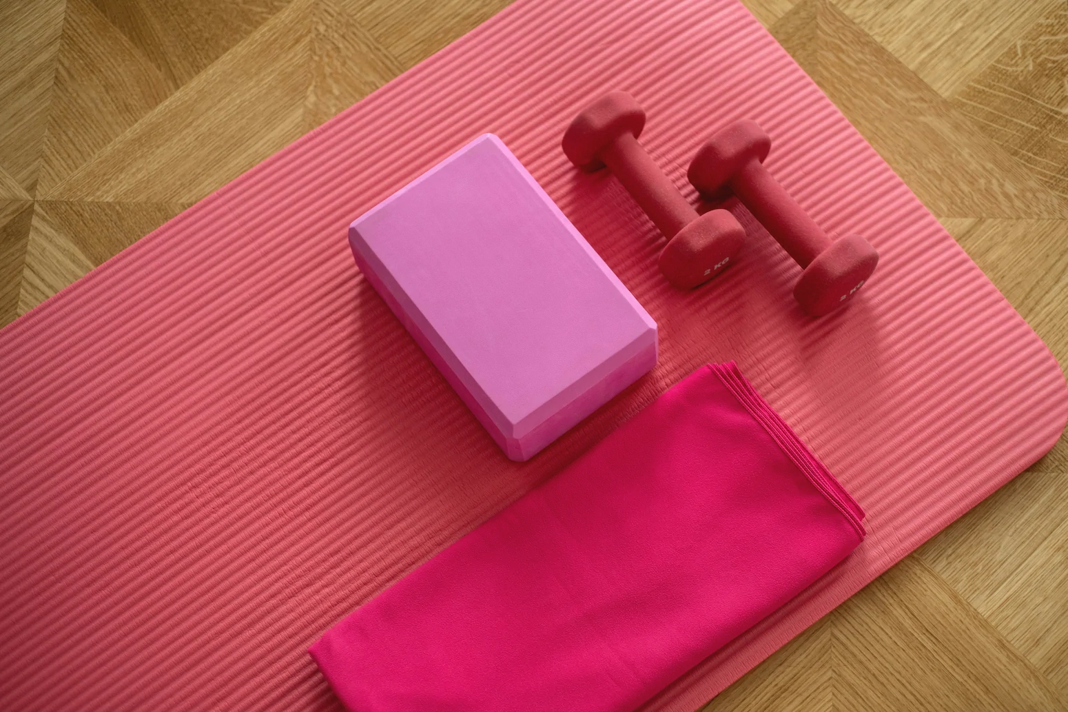 A pink exercise mat with a yoga block, two red dumbbells, and a folded pink towel on a wooden floor. Ohio