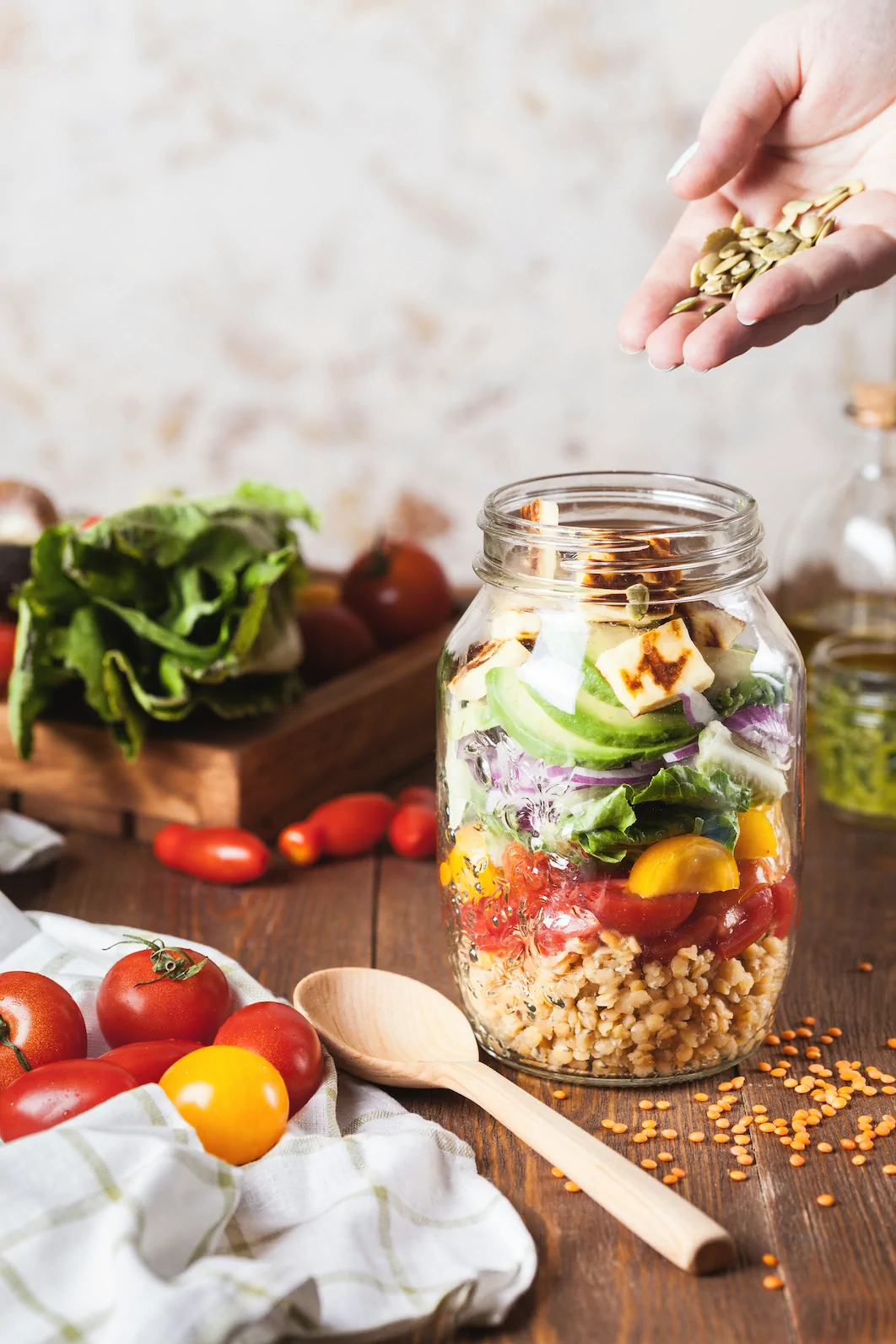 A glass jar filled with layers of grains, chopped vegetables, and cheese; a hand sprinkles seeds on top. Fresh vegetables and a wooden spoon are on the table nearby. Ohio